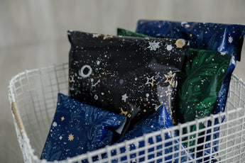 A white wire basket contains several wrapped packages in Midnight Galaxy Mailers by impack.co, showcasing starry and leafy designs in blue, black, and green hues for a stylish look while ensuring safe transit.