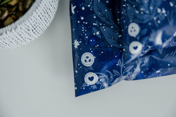 The Midnight Indigo Mailers (10" x 13") by impack.co feature blue patterned packaging with white symbols, like circles and hearts, symbolizing sustainability. A textured white bowl at the top left reinforces their commitment to eco-friendly packaging without sacrificing quality.