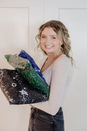 A person smiling while holding three wrapped gifts in impack.co's SALE - Midnight Galaxy Mailers 14.5" x 19" against a light background.