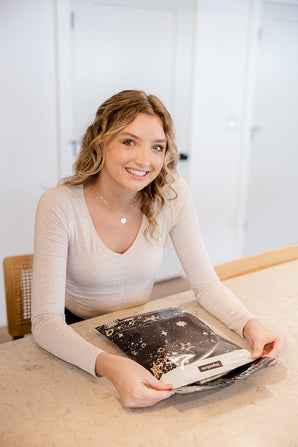 A woman sitting at a kitchen counter holding a book, surrounded by impack.co's Midnight Galaxy Mailers 10" x 13", zero waste and recyclable products.