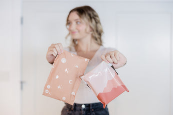 A person holds two decorative packages, one with celestial patterns and the other featuring a mountain design from impack.co's Mountain Clay Mailers 10" x 13", highlighting the use of biodegradable mailers for transit protection against a simple backdrop.