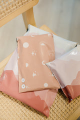 Three biodegradable Celestial Tan mailers by impack.co, sized at 6" x 9", sit stylishly on a woven chair, providing both vibrant patterning and transit protection.