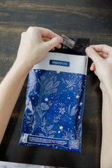 Hands opening a package with a blue floral pattern labeled "impack.co" on a dark wooden table, featuring the SALE - Midnight Indigo Mailers 6" x 9", offering durable and sustainable biodegradable protection.