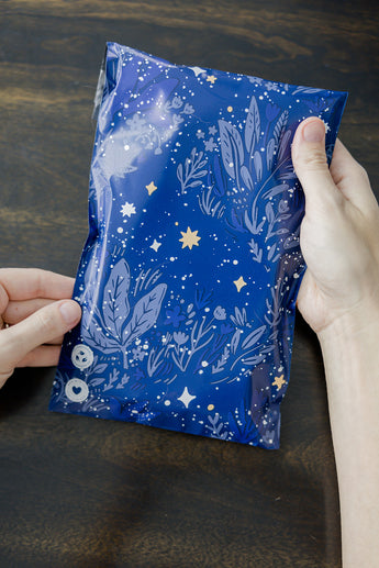Hands display a SALE - Midnight Indigo Mailer by impack.co, adorned with blue floral and star designs, on a dark wooden surface, symbolizing robust protection and sustainability.