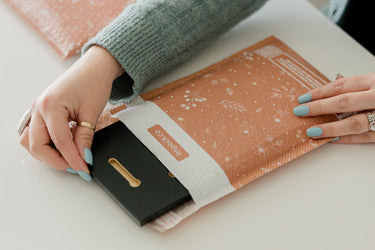 Hands with blue nails placing a black item into a decorative brown padded envelope.