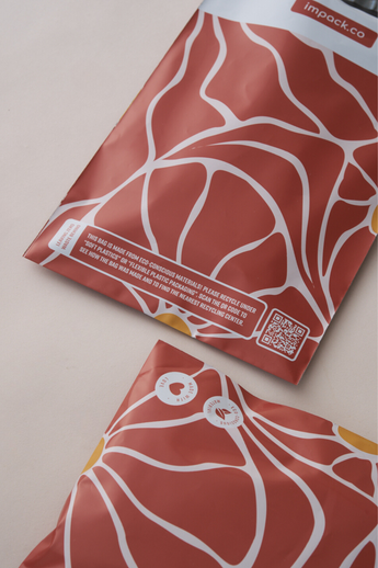 Close-up of two "SALE - Crimson Grove Mailers 10\" x 13\"" by impack.co, featuring red and white patterns with eco-friendly messages and a QR code on a beige surface. These biodegradable mailers offer superior transit protection while being environmentally friendly.