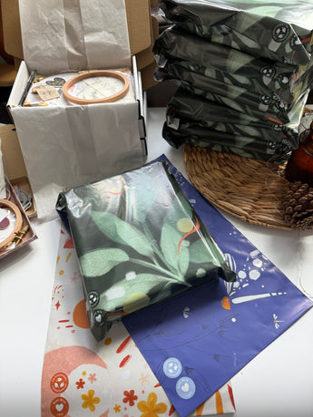 A stack of gift-wrapped packages with green leaf patterns sits beside impack.co’s Boho Sun & Moon Biodegradable Mailers (14.5" x 19"), decorated envelopes, boho packaging, and assorted craft supplies on a table.