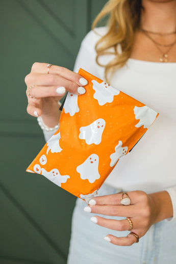 A person holds an impack.co SALE- Halloween Ghost Pumpkin Biodegradable Mailer (6" x 9", CA) featuring white cartoon ghosts; only their torso and hands are visible.