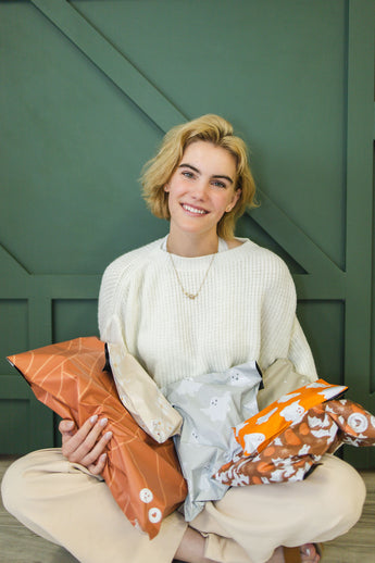 A person with short blonde hair in a white sweater sits cross-legged, holding impack.co's SALE-Halloween Ghost Pumpkin Biodegradable Mailers (6" x 9") against a green background.
