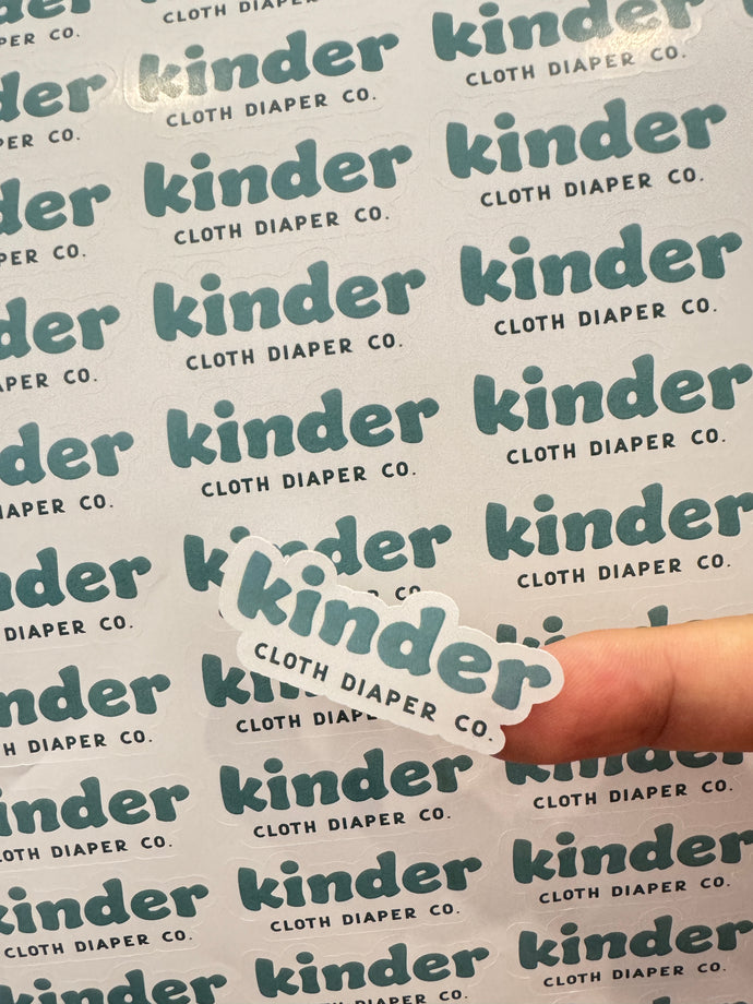 A hand holds a sticker featuring the logo "kinder Cloth Diaper Co." against a background covered with the same logo.
