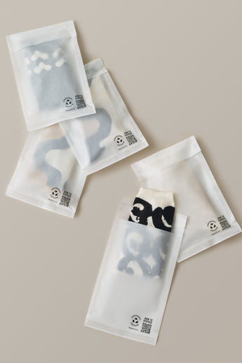 Five semi-transparent Glassine Bags 5.6" x 8.6" from impack.co contain folded clothing with different patterns, placed on a neutral background. Each bag has printed text and a QR code.