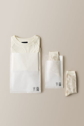 A folded white shirt and a pair of cream patterned socks are neatly packaged in impack.co Glassine Bags 9.6" x 12.6", with an additional single sock placed outside the bag.