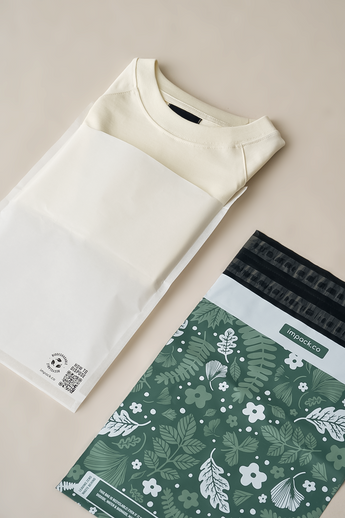 A folded beige shirt lies next to a green, floral-patterned Glassine Bags 9.6" x 12.6" by impack.co on a beige surface.