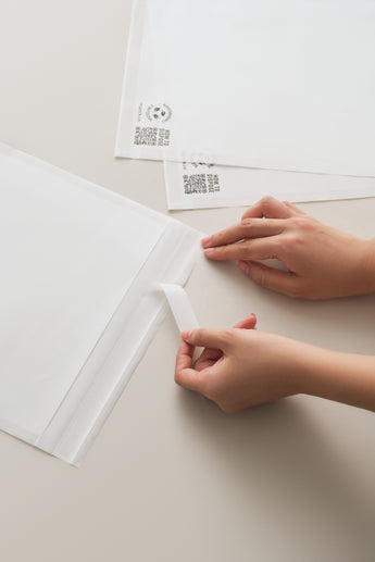 Hands peeling a strip from impack.co Glassine Bags 9.6" x 12.6" on a light-colored surface. Two more glassine bags and barcode labels are visible in the background.