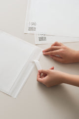 Hands peeling a strip from impack.co Glassine Bags 9.6" x 12.6" on a light-colored surface. Two more glassine bags and barcode labels are visible in the background.