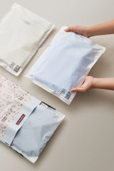 Hands holding a sealed translucent impack.co Glassine Bag 9.6" x 12.6" containing a folded garment, with three similar bags lying on a light surface nearby.