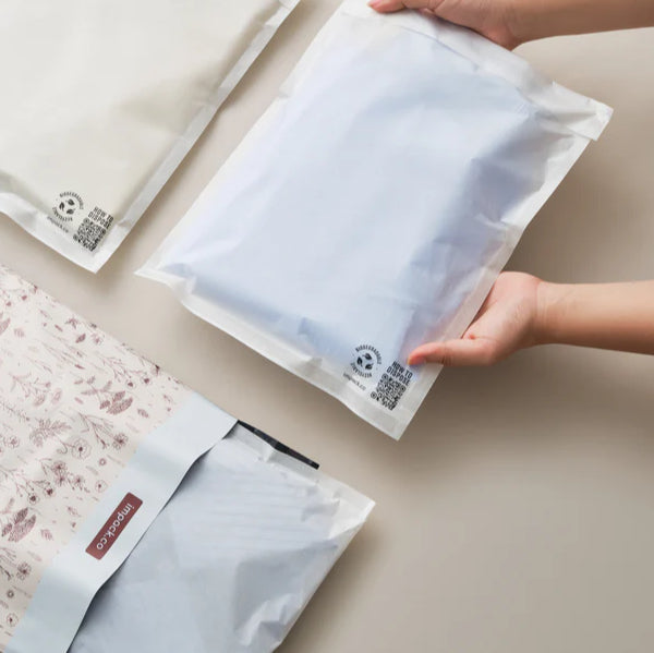 A person holds a semi-transparent, sealed plastic bag containing folded fabric; two similar bags and a floral-patterned pouch are on a light surface.
