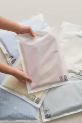 Hands are placing a folded pink t-shirt into a translucent resealable bag. Several other sealed bags each containing different colored clothing items are spread out on a light surface.

Adjusted Sentence: Hands are placing a folded pink t-shirt into an impack.co Glassine Bag 9.6" x 12.6". Several other sealed impack.co Glassine Bags, each containing different colored clothing items, are spread out on a light surface.