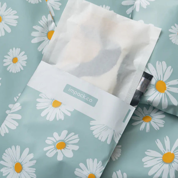 A light blue mailing bag with white and yellow daisy prints, partially open to reveal a white paper package and label with "impack.co" printed on it.