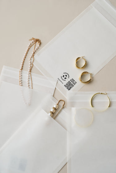 Several impack.co Glassine Bags 5.6" x 8.6" containing golden hoop earrings and pearl necklaces are laid out neatly on a beige surface.