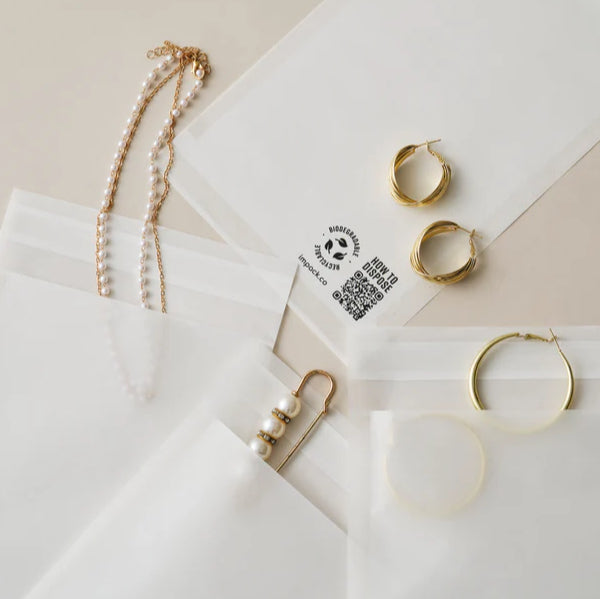 Several pieces of gold jewelry, including necklaces and hoop earrings, are placed on and in translucent white pouches with recycling symbols and QR codes printed on them.