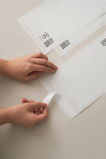 A person peeling a strip from an impack.co Glassine Bags 5.6" x 8.6", placed on a light beige surface.