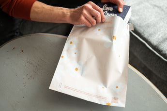 Someone gently places a book into an impack.co SALE - Cosmos Biodegradable Mailer (10" x 13"), featuring a white floral pattern on a table, ensuring its transit protection.