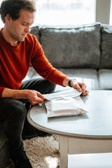 A man in a red sweater carefully opens an impack.co Cosmos Biodegradable Mailer (10" x 13" CA) on a round white table. These mailers offer great transit protection and neatly align with his eco-friendly lifestyle.