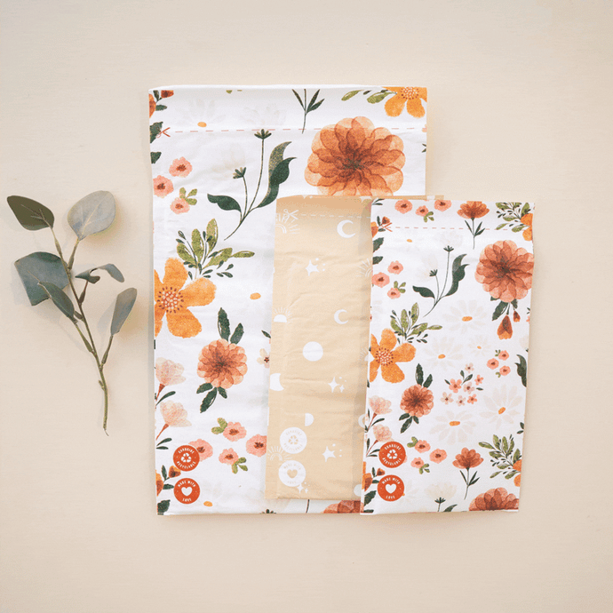 Flat lay of folded floral-patterned bedsheets with orange and white flowers, placed next to a small sprig of green leaves on a beige background.