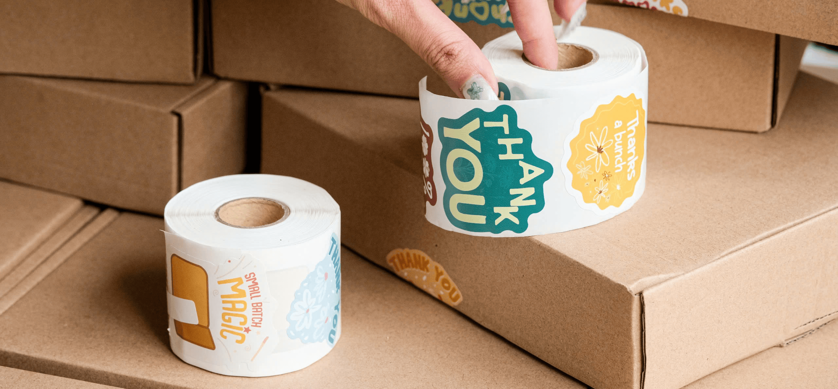 A hand holds a roll of colorful thank you stickers above stacked cardboard boxes, with another sticker roll and applied stickers visible.