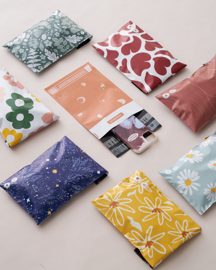 Nine colorful mailer bags with various floral, celestial, and abstract patterns arranged in a grid on a light surface, with one bag partially open showing contents inside.