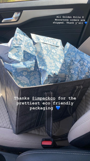 A car seat with a large gray tote bag holding multiple blue, floral-patterned packages. Overlay text reads: "All Golden Hills X Moonstone orders are shipped. Thank y'all. Thanks @impack.co for the prettiest eco-friendly packaging and robust protection using Sapphire Evergreen Biodegradable Mailers 6" x 9".
