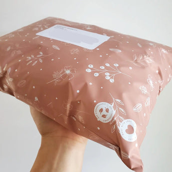 A hand holds impack.co’s SALE - Floral 2D Rosy Brown Biodegradable Mailer (10" x 13"), featuring a floral pattern, shipping label, and eco-friendly symbols for stylish and reliable package protection.