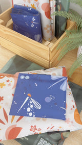 Boho Sun & Moon Biodegradable Mailers (14.5" x 19") by impack.co—featuring colorful, boho-inspired designs—are displayed in a wooden box and on a table beside a green fern plant.