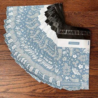A stack of Sapphire Evergreen Biodegradable Mailers 6" x 9" by impack.co, recyclable blue and white envelopes, on a wooden floor.