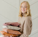 A person with blonde hair, wearing a beige top, holds a stack of folded patterned clothes featuring the Natural Cedar Leaf Biodegradable Mailers by impack.co against a light-colored wall.