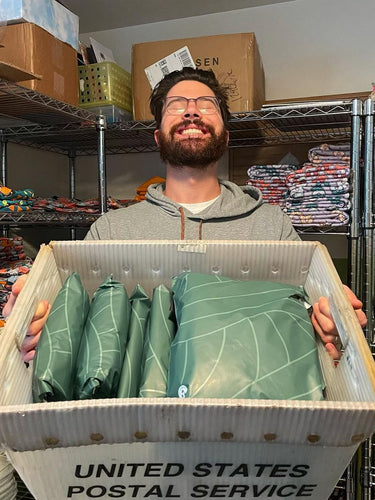 Person smiling while holding a large USPS box filled with green wrapped packages, standing in a room with shelves of colorful fabric.