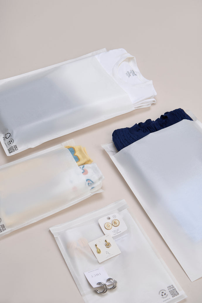 Folded shirts in t-shirt packaging, pants, socks, and accessories like earrings and rings are neatly arranged on a flat surface. The display also includes eco-friendly Glassine Bags (14.1" x 18.6") by impack.co, adding a touch of sustainability with their translucent packaging.