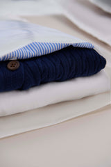 Folded clothes, such as a blue knitted sweater with a button and a white shirt with blue stripes, rest on a light surface. Ready for travel, they sit comfortably in eco-friendly Glassine Bags 14.1" x 18.6" by impack.co, ideal for packaging and shipping.