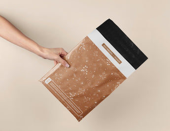 A hand holds impack.co’s SALE - Floral 2D Rosy Brown Biodegradable Mailer (10" x 13") with a black adhesive strip, highlighting eco-friendly, stylish packaging ideal for secure shipping on a beige background.