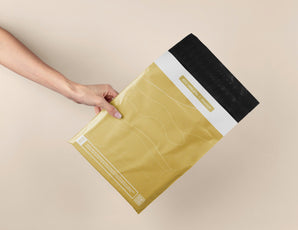 A hand displays the impack.co SALE - Tidal Amber Mailers (10" x 13") in gold and black on a beige background, highlighting their eco-friendly design suitable for medium-sized shipments.