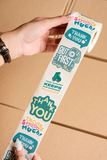 A person holds an impack.co Sticker Roll featuring colorful packaging stickers with phrases like “Thank you” and “But first snap me” over a brown box, perfect for adding fun to any package.