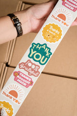 A hand holds an impack.co Sticker Roll featuring colorful eco-friendly stickers with gratitude messages like "Thank You," "Thanks a bunch," and "100% Grateful" over packed boxes—ideal for packaging.