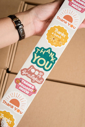 A hand holds an impack.co Sticker Roll featuring colorful eco-friendly stickers with gratitude messages like "Thank You," "Thanks a bunch," and "100% Grateful" over packed boxes—ideal for packaging.
