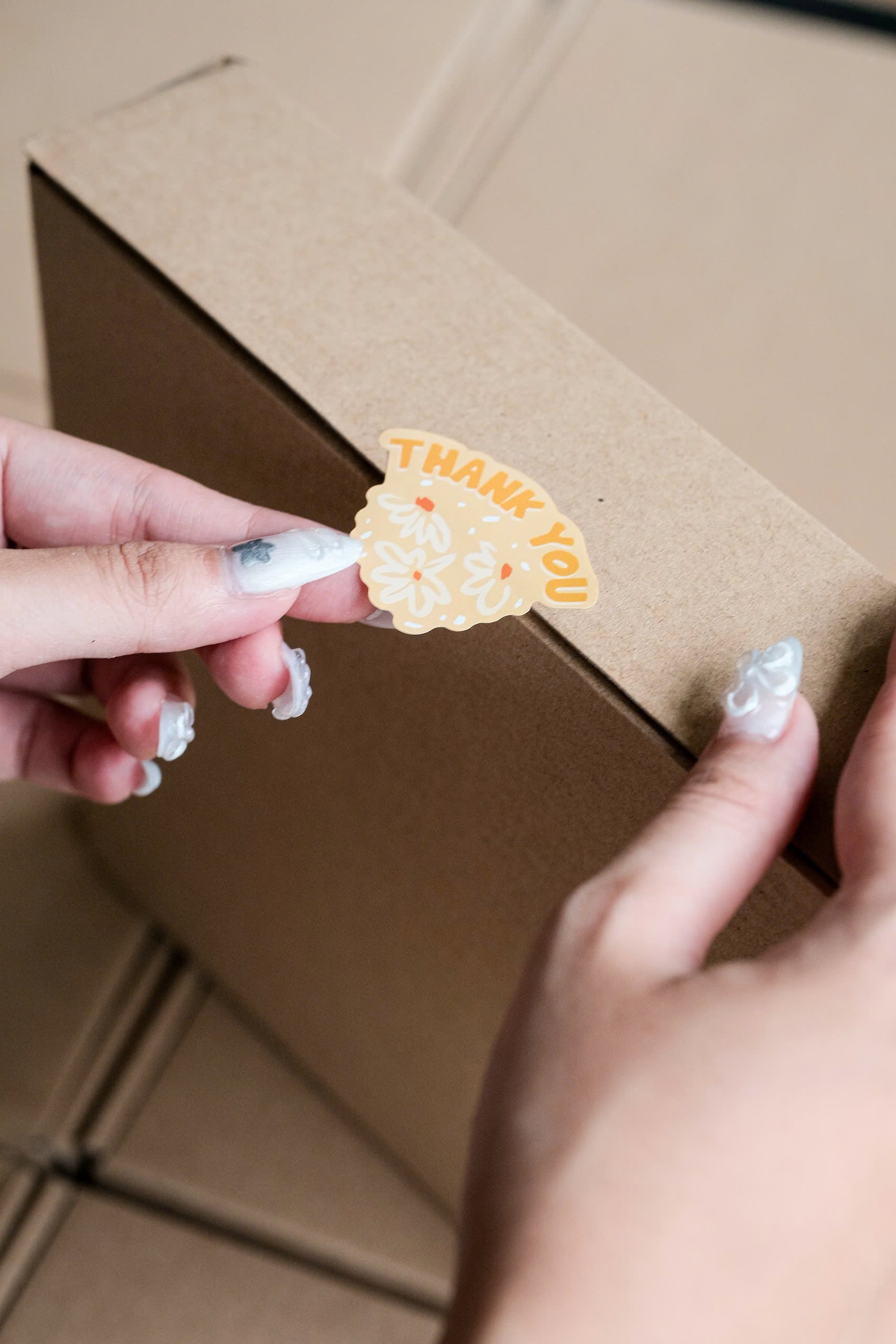 A person applies an eco-friendly impack.co Sticker roll 500 pcs with yellow "Thank You" floral packaging stickers onto a closed cardboard box.