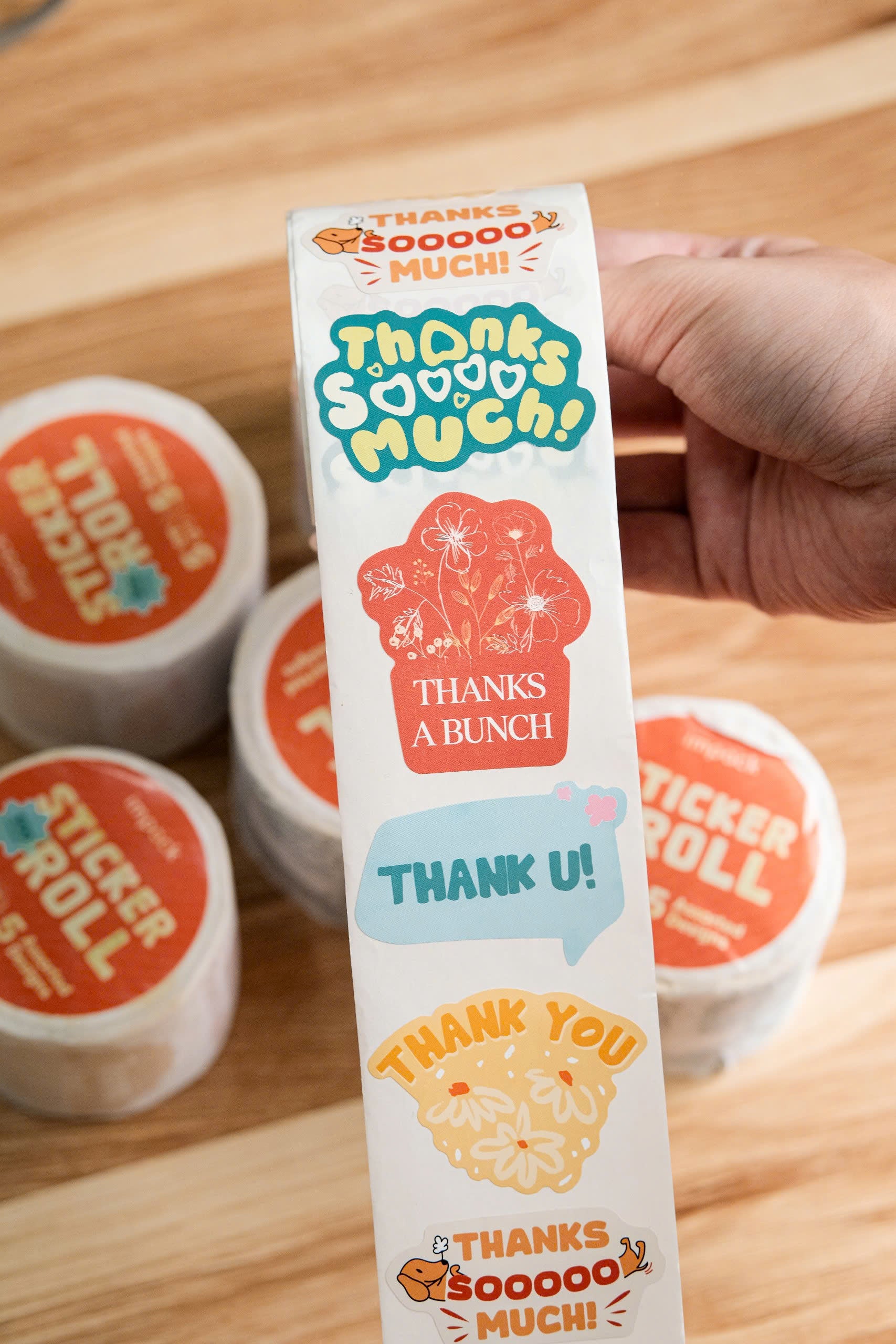 A hand holds a roll of colorful stickers with messages like "Thanks so much!", "Thanks a bunch", "Thank you", and "Thank u!" above sticker rolls on a wooden surface.