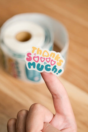 A hand displays a vibrant "Thanks so much!" sticker from the impack.co Sticker Roll, made with recyclable paper, as a roll of eco-friendly stickers lies blurred on a wooden surface in the background.