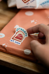 A hand places an impack.co Sticker Roll, which is a recyclable paper sticker reading "Spreading love at 2AM," on peach-colored gift wrap with white polka dots.
