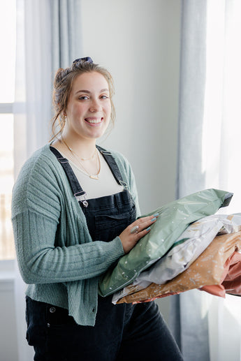 A person in a green sweater and dark overalls stands indoors near a window with light curtains, holding several SALE - Starry Lily Pad Biodegradable Mailers 14.5" x 19" by impack.co, carefully packed for transit protection.