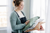 A woman in a sweater and overalls holds several colorful, patterned impack.co SALE - Starry Lily Pad Biodegradable Mailers 14.5" x 19" by the window, ready for transit protection.